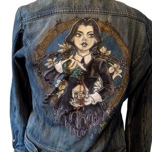 以下が含まれることがあります： 背面に手描きデザインが施された青いデニムジャケット。デザインは、黒いドレスを着た女性が血が滴る頭蓋骨を持っている様子を描いています。デザインは金色の枠と花のアクセントで囲まれています。