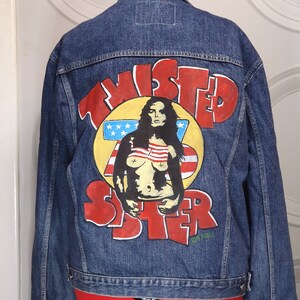 Op de afbeelding: Een blauwe denimjas met een handgeschilderd ontwerp op de rug. Het ontwerp bevat de woorden "Twisted Sister" in rode en gele letters, met een vrouw in een rood-wit-blauwe outfit die voor een Amerikaanse vlag staat.
