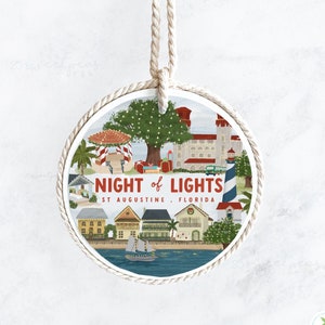 May include: A round, white ornament with a rope hanger. The ornament features a colorful illustration of a cityscape with the text "NIGHT of LIGHTS ST AUGUSTINE, FLORIDA".