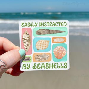 May include: A square sticker featuring the text EASILY DISTRACTED BY SEASHELLS in green letters. The design includes six illustrations of various seashells in shades of brown, tan, and pink, set against colorful rectangular backgrounds.