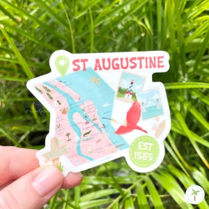 May include: A pink and blue illustrated map sticker of St. Augustine, Florida. The map shows the city's landmarks and includes a red bird, a lighthouse, and a beach. The sticker also includes the text "EST 1565".