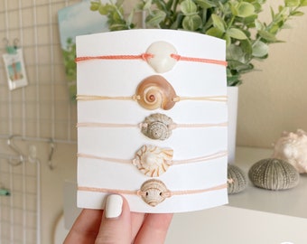 May include: Five bracelets with different seashells attached to pink and beige cords. The bracelets are displayed on a white card.