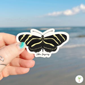 May include: A sticker featuring a Zebra Longwing butterfly. The butterfly has black wings with yellow stripes and a white border. The text "Zebra Longwing" is below the butterfly. The background is a beach scene with blue water and sky.