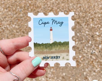 May include: A rectangular sticker shaped like a postage stamp featuring an illustration of the Cape May lighthouse in New Jersey. The design includes a white lighthouse with a red top, green dunes, a wooden fence, and blue sky with text reading Cape May.