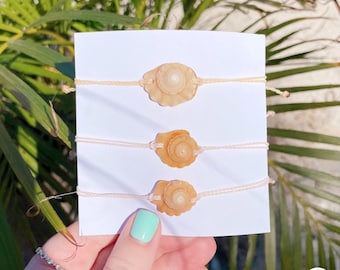 May include: Three shell bracelets on a white card. The bracelets are made with natural shells and white string. The shells are different sizes and shapes.