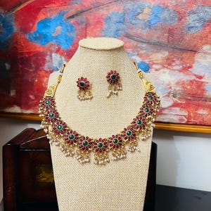 May include: A gold-coloured necklace and matching earrings set. The necklace and earrings feature a floral design with red and green gemstones, accented with small white pearls. The jewellery is displayed on a beige bust.
