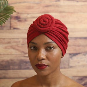 May include: A red turban with a spiral knot detail. The head covering is made of a soft fabric and is worn by a person. The background is a wooden wall with a green plant.