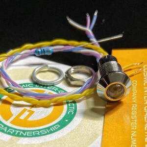 May include: A chrome-plated LED indicator light with a yellow lens. The light is surrounded by a bundle of wires, including a blue resistor. The wires are connected to a small metal nut and a metal ring.