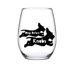 May include: A clear stemless wine glass with a black silhouette of two running dogs. The words "Dogs Before Knobs" are printed in black on the glass, creating a fun and playful design. Perfect for dog lovers.
