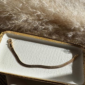 May include: A gold-colored, flat snake chain bracelet rests on a white tray with a gold rim. The bracelet is made of small, interlocking links. The background features a soft, textured, beige material.