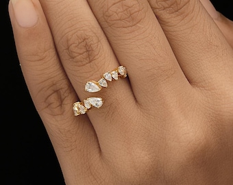 May include: A gold-colored ring with a wrap-around design, adorned with multiple pear-shaped and round clear gemstones. The ring is displayed on a hand against a black background, showcasing its delicate details and sparkle.