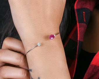May include: A silver bangle bracelet with a small pink stone and a clear stone. The bracelet is on a person's wrist.