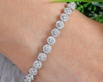 Puede incluir: Una pulsera plateada con numerosas piedras preciosas redondas y transparentes. La pulsera se lleva en una muñeca, con una manga verde y flores en el fondo. Las piedras están muy juntas.