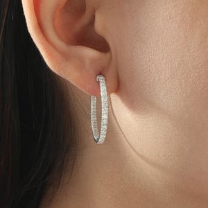 May include: A close-up of a silver hoop earring with small, clear stones set along the outer surface. The earring is worn on the ear of a person with dark hair. The earring is a classic, elegant jewelry piece.