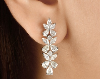 May include: A close-up of a dangling earring with a floral design. The earring features multiple clear, sparkling gemstones arranged in a cascading pattern. The gemstones are set in a silver or white metal.