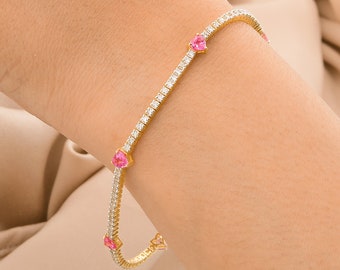 Puede incluir: Una pulsera dorada con un diseño delicado. La pulsera presenta pequeñas piedras claras y brillantes y gemas rosas en forma de corazón espaciadas uniformemente. La pulsera se muestra sobre un fondo de tela beige.