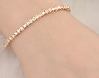 Puede incluir: Una pulsera de tenis dorada con una fila de piedras preciosas claras, de corte cuadrado. La pulsera se lleva en una muñeca, con un fondo blanco suave y detalles florales. Las piedras preciosas brillan.
