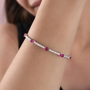 Pode incluir: Uma pulseira de metal prateado com uma fila contínua de pequenas pedras transparentes de corte redondo, intercaladas com quatro gemas maiores de corte oval num vibrante tom vermelho-rosado, dispostas a intervalos regulares.