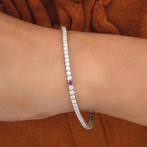 Könnte beinhalten: Ein filigranes Silberarmband, besetzt mit zahlreichen kleinen, klaren Edelsteinen. Ein einzelner, leuchtend roter Edelstein ist in die Reihe der klaren Steine eingefügt. Das Armband wird auf einem Arm vor warmbraunem Hintergrund gezeigt.