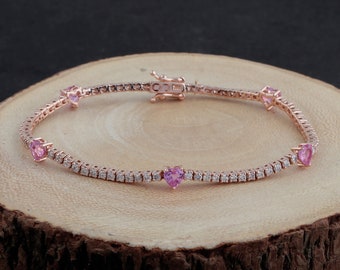 May include: Rose gold bracelet featuring a row of small, clear stones and heart-shaped pink gemstones. The bracelet is set on a wooden surface, showcasing its delicate design and sparkle. A beautiful piece of jewelry.