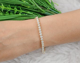 Puede incluir: Una pulsera de tenis dorada con una fila de piedras preciosas claras y brillantes. La pulsera se muestra en una muñeca, con follaje verde y una superficie blanca y texturizada en el fondo.