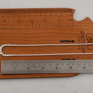 May include: A delicate silver necklace with clear, round gemstones, displayed on a wooden ruler. The necklace is approximately 18 inches long. The ruler shows measurements in inches, and the necklace is laid out for display.