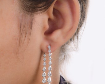 May include: Silver-toned drop earrings featuring multiple clear, pear-shaped gemstones. The earrings cascade downwards, creating a sparkling effect. The earrings are worn on a person's ear.