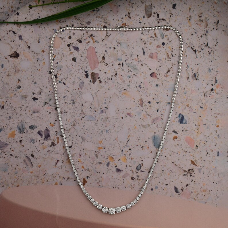 May include: A delicate, silver-toned necklace with numerous small, sparkling stones. The necklace features a graduated design, with larger stones clustered at the center and smaller stones extending along the chain. The necklace is displayed on a speckled, neutral-toned surface.