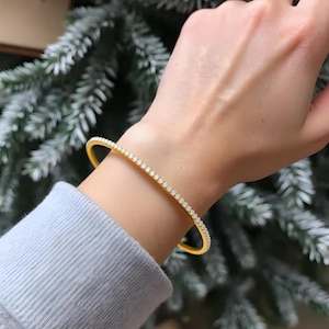 May include: A gold-colored bracelet with a row of small, clear gemstones. The bracelet is worn on a wrist, and the background is a blurred view of a green and white Christmas tree.