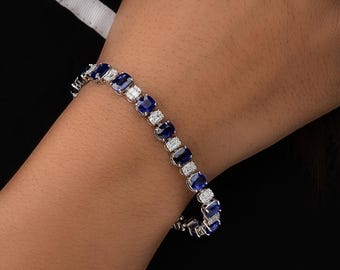 May include: A silver bracelet featuring alternating oval blue gemstones and rectangular clear gemstones. The bracelet is displayed against a dark background, showcasing the sparkle of the gems. The bracelet is a piece of jewelry.