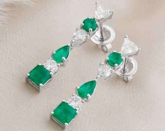 May include: Drop earrings with emerald and diamond gemstones. The earrings have a linear design with a square emerald, a diamond, a pear-shaped emerald, another diamond, a round emerald, and a triangular diamond stud. The earrings are set in a silver metal.