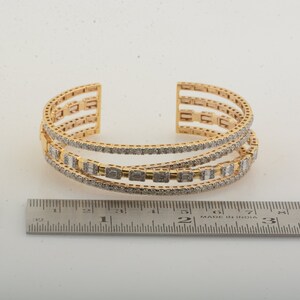 May include: Gold-toned cuff bracelet with three rows of rectangular and round-cut gemstones. The open design features multiple parallel bands, with a ruler in the foreground. The bracelet is likely made of gold and gemstones.