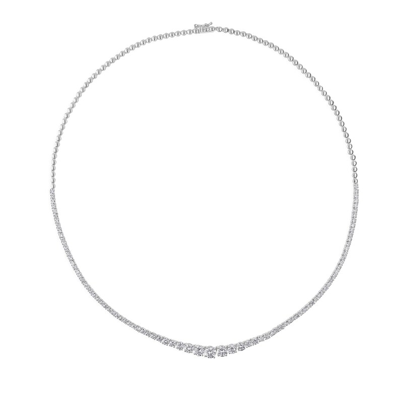 May include: A delicate, silver-toned necklace with a continuous row of small, round, clear gemstones. The necklace curves gracefully, with a slight dip in the center, and is secured with a small clasp. The gemstones sparkle against a white background.