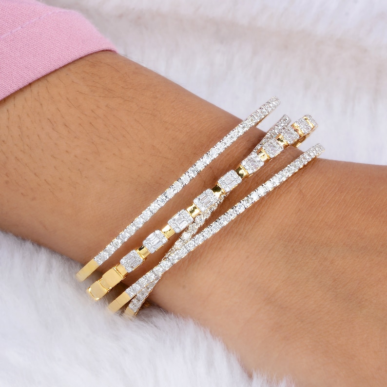 May include: Four gold and diamond bracelets are stacked on a wrist. The bracelets feature a mix of round and baguette-cut diamonds set in gold bands. The diamonds sparkle against the gold, creating a luxurious and elegant look. The bracelets are displayed on a white, fluffy surface.