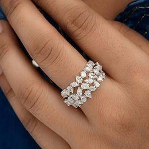 Puede incluir: Primer plano de un anillo de diamantes con múltiples piedras en forma de pera engastadas en una banda. El anillo es plateado y brilla, reflejando la luz. El anillo está en el dedo de una persona, con un fondo azul.