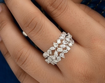 May include: A close-up of a diamond ring featuring multiple pear-shaped stones set in a band. The ring is silver-toned and sparkles, reflecting light. The ring is on a person's finger, with a blue background.