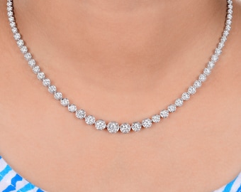 May include: A diamond necklace with multiple round-cut diamonds set in a white metal. The necklace is draped around a person's neck, showcasing the graduated size of the diamonds. The background is a light blue and white striped pattern.