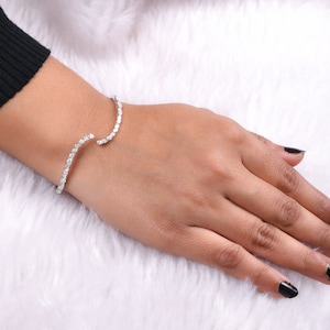 May include: A silver-toned open cuff bracelet with a row of small, clear gemstones. The bracelet curves around the wrist, with the gemstones set along the outer edge. The bracelet is on a hand with black nail polish.