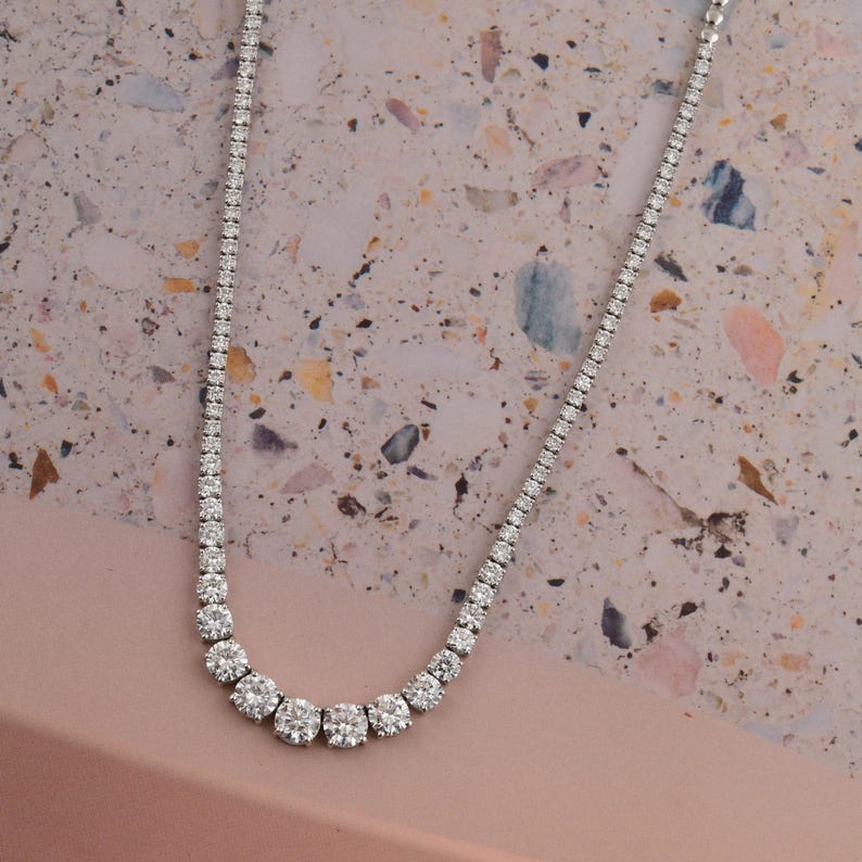 May include: A delicate, silver-toned necklace with a graduated design. The necklace features a series of round, sparkling stones that increase in size towards the center. The stones are set closely together, creating a continuous line of brilliance. The necklace is displayed on a pink and beige surface.