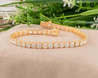 Puede incluir: Una pulsera de tenis dorada con piedras preciosas claras de talla cuadrada. La pulsera está colocada sobre una superficie de color neutro, con follaje verde en el fondo. El cierre es visible y la pulsera parece nueva.