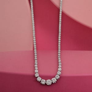 May include: A diamond tennis necklace with a graduated design. The necklace features a series of round, brilliant-cut diamonds set in a white metal. The diamonds increase in size towards the center, creating a sparkling, elegant look. The background is a gradient of pink hues.