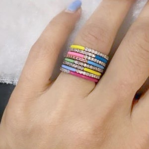 Può includere: Una pila di anelli smaltati colorati con una fila di piccole pietre trasparenti. Gli anelli sono nelle tonalità di rosa, giallo, blu, verde e azzurro. Gli anelli sono su un dito con un'unghia azzurra.