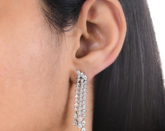 May include: Silver drop earrings featuring multiple rows of clear, sparkling stones. The earrings have a cluster of stones at the top and a cascading design, creating a glamorous and elegant look. The earrings are shown on a person's ear.