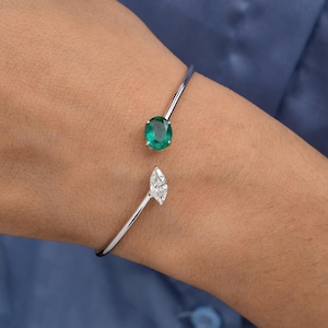 May include: A silver-toned cuff bracelet featuring an oval emerald and a marquise-cut diamond. The bracelet's open design allows it to wrap around the wrist. The emerald is a vibrant green, and the diamond is clear and sparkling.