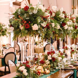 Puede incluir: Centros de mesa florales con rosas rojas y blancas, vegetación y bayas, dispuestos sobre altos soportes dorados. La mesa incluye velas, cristalería y un camino de mesa verde. Ancho 51 cm.