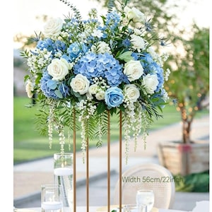 Peut inclure: Centre de table floral avec des roses blanches, des hortensias bleus et de la verdure. Le support est en métal doré, largeur 56 cm. La table est ornée de bougies et de verres.