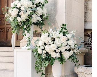 May include: Two white urns filled with white flowers and greenery, each atop a white pedestal. The urns are 22 inches (55 cm) wide. The arrangement is set against a stone building with a wooden door.