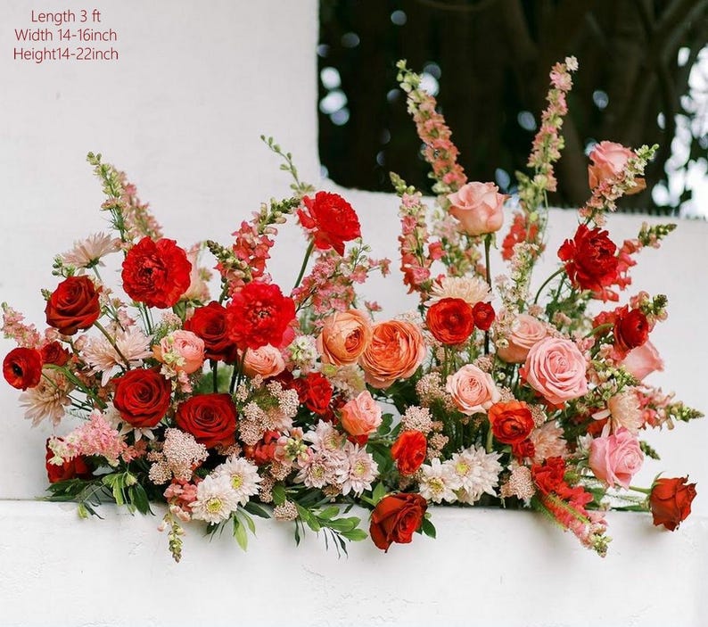 May include: A vibrant floral arrangement featuring a variety of blooms in shades of red, pink, and peach. The arrangement is approximately 3 feet long, 14-16 inches wide, and 14-22 inches high. Roses, carnations, and other flowers create a lush display.