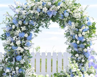 Puede incluir: Un arco floral, de aproximadamente 244 cm de ancho y 244 cm de alto, adornado con flores azules y blancas. El arco se encuentra frente a un cielo azul y una valla blanca. El texto "ArchFlowers W8xH8 ft" es visible.