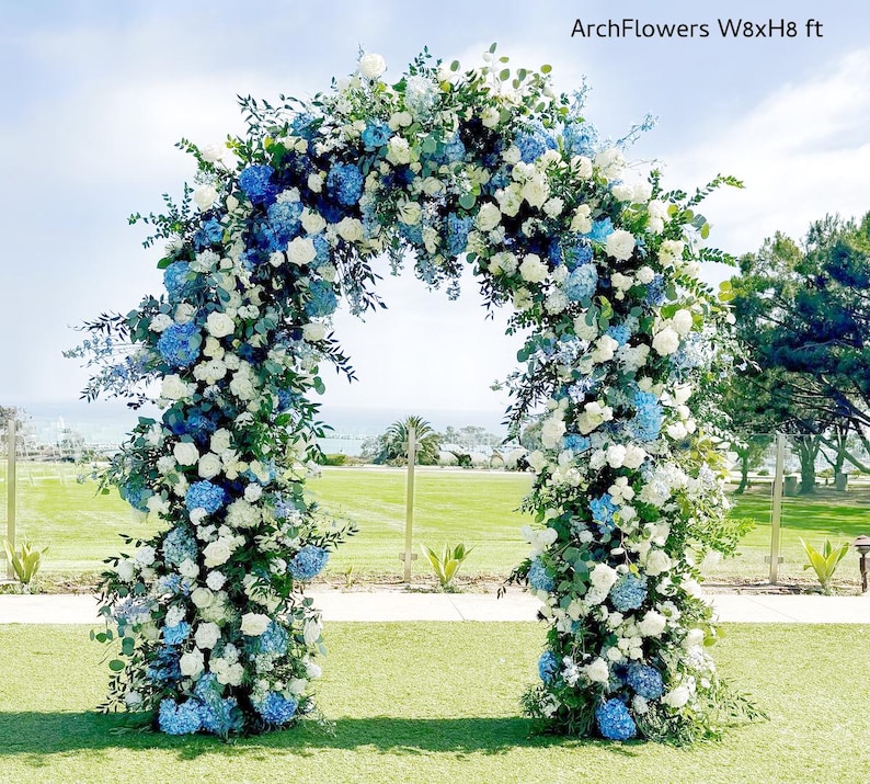May include: A floral archway, 8 feet wide and 8 feet high, adorned with white roses, blue hydrangeas, and greenery. The arch is set on a green lawn with a blue sky background. Text on the image reads "ArchFlowers W8xH8 ft".